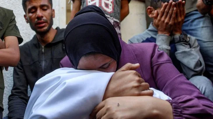 امرأة تحتضن طفلاً، بينما يظهر في الخلفية رجال يظهر عليهم الحزن، في سياق الهجمات الإسرائيلية على غزة التي أودت بحياة العديد من الفلسطينيين.