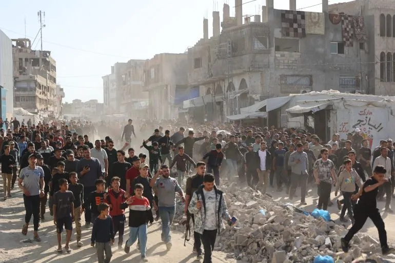 محتجون يسيرون في شارع غزّي مغطى بالأنقاض، وسط غبار كثيف، مع وجود مبانٍ مدمرة في الخلفية، يعكس تصاعد العنف في المنطقة.