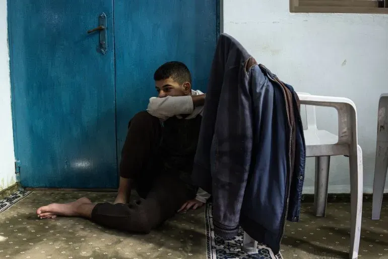 شاب فلسطيني يجلس على الأرض بجوار باب أزرق، يعبر عن الإحباط والقلق بعد اعتقاله، وسط أجواء من التوتر في طوباس.