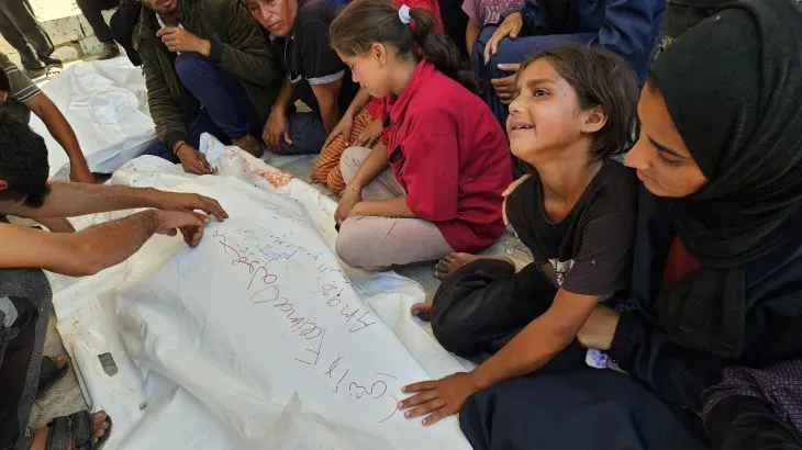 أطفال فلسطينيون يجلسون بجانب جثث على الأرض، مع تعبيرات حزن على وجوههم، في سياق الأزمة الإنسانية المتزايدة في غزة.