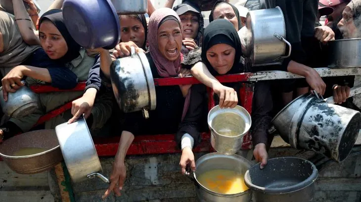 نساء وأطفال فلسطينيون يتجمعون بقلق حول نقطة توزيع الطعام في غزة، مع أواني معدنية، في ظل أزمة إنسانية متفاقمة.
