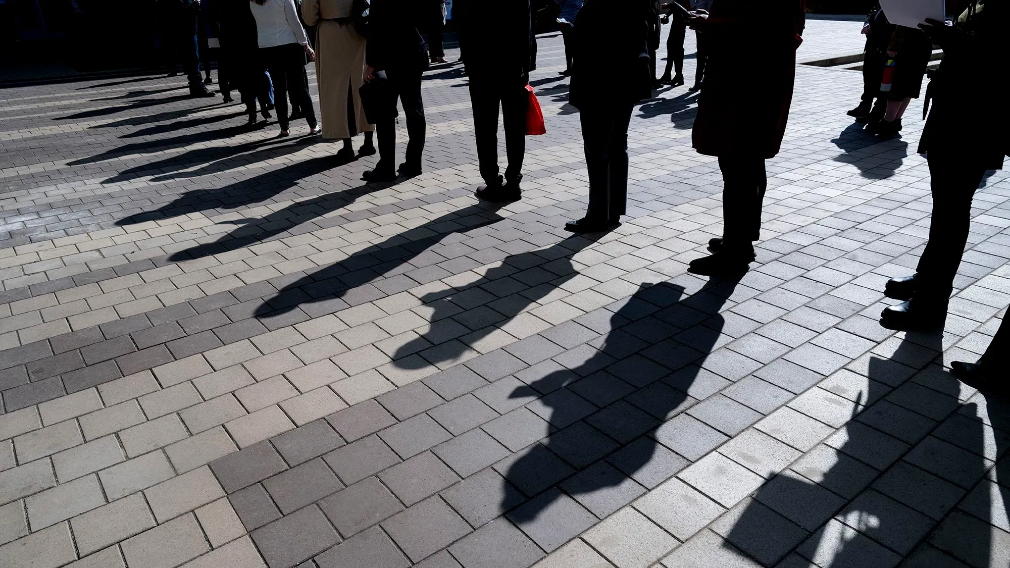 طابور من الأشخاص يقفون على رصيف مكون من بلاط ملون، مما يعكس حالة عدم اليقين في سوق العمل وارتفاع طلبات إعانات البطالة.