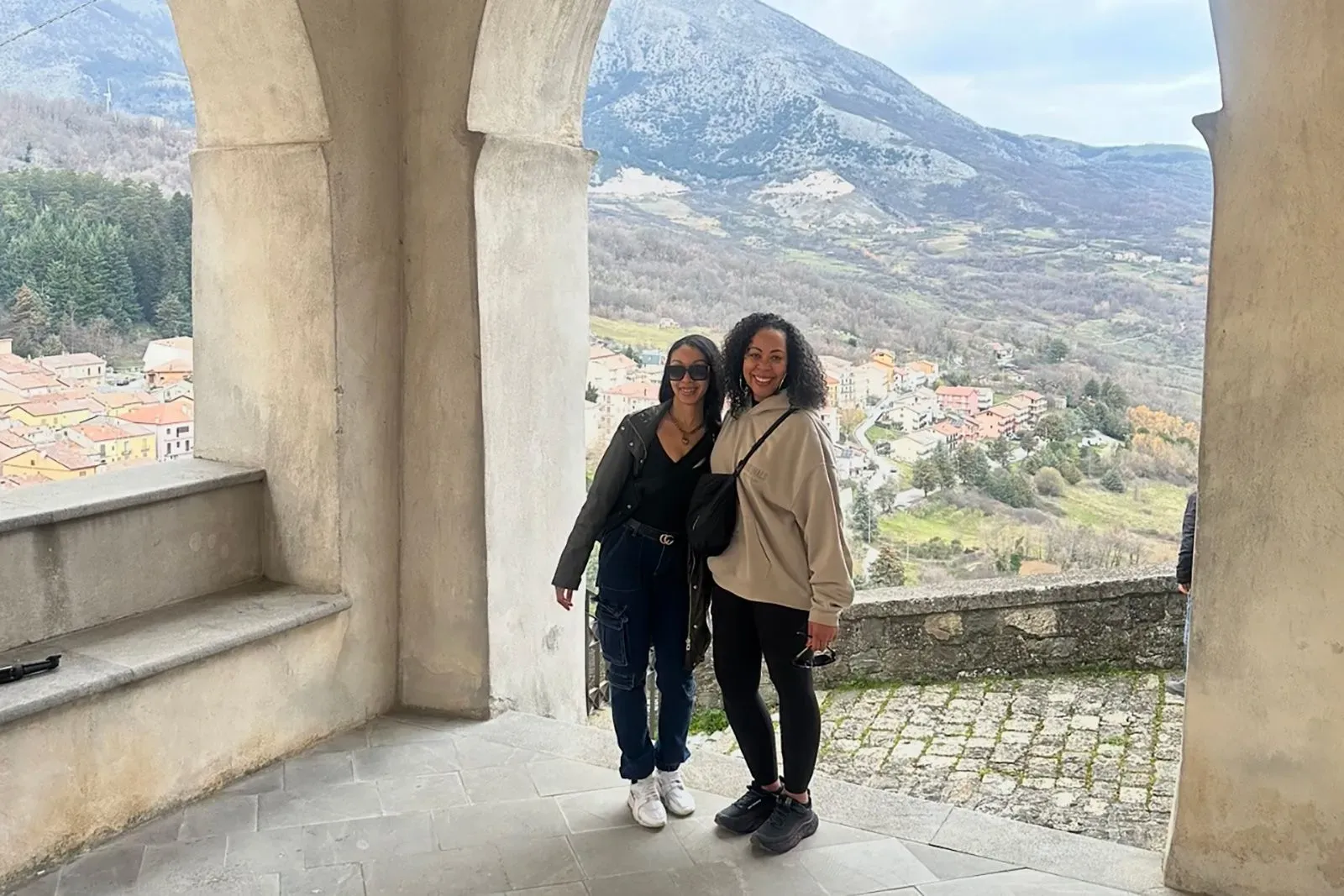 امرأتان تبتسمان معًا في موقع مرتفع يطل على قرية لاترونيكو الإيطالية، حيث تظهر المناظر الطبيعية والجبال المحيطة.