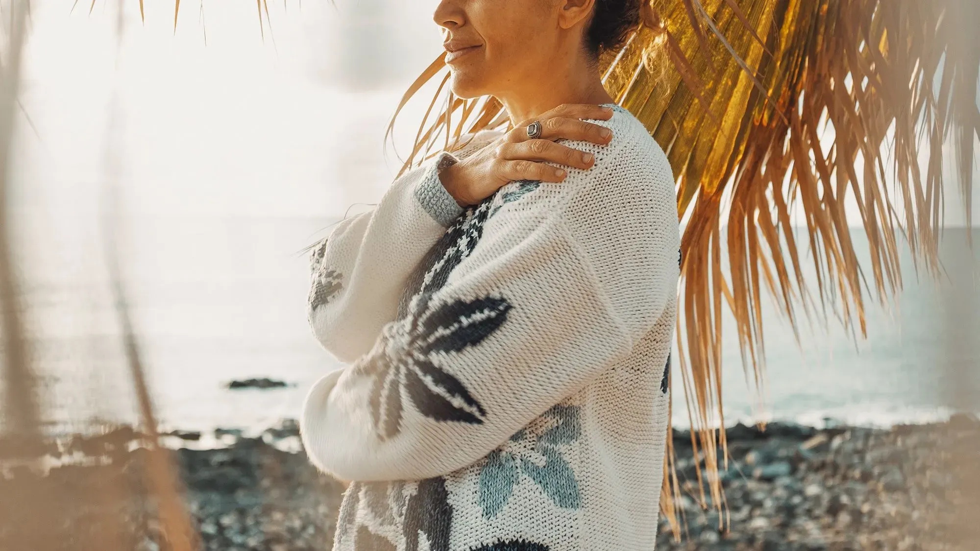 امرأة تعانق نفسها برفق على شاطئ، تعبيرها يوحي بالهدوء والقبول الذاتي، مما يعكس موضوع إعادة التربية والتعاطف مع النفس.