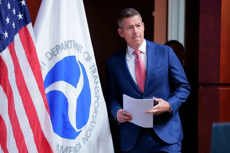 رجل يرتدي بدلة رسمية يقف بجانب علم وزارة النقل الأمريكية، يبدو جادًا أثناء مغادرته قاعة مؤتمرات.