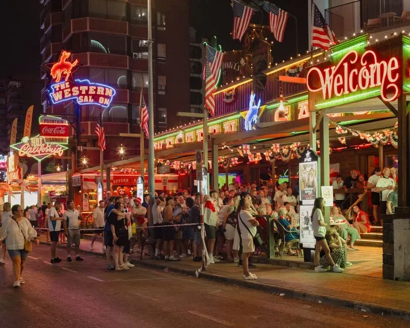 مشهد لشارع مزدحم في بينيدورم ليلاً، حيث تتلألأ الأضواء الملونة لواجهات الحانات والمطاعم، مع تجمع السياح للاستمتاع بأجواء المدينة.