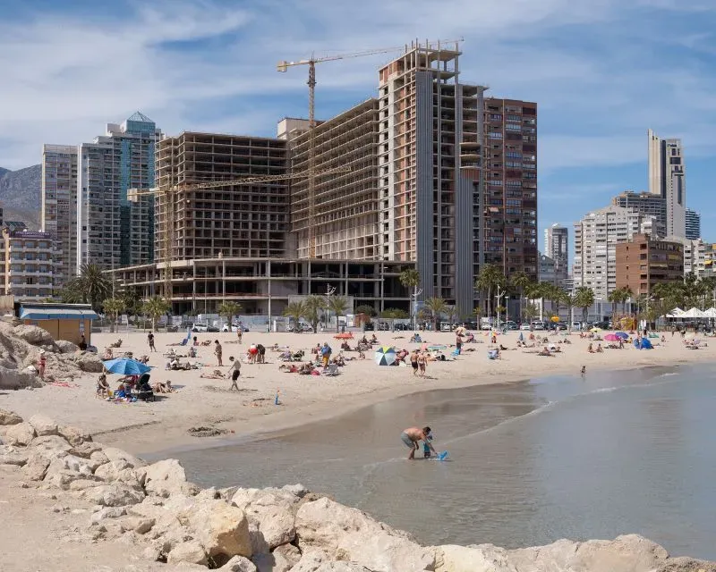 شاطئ بينيدورم مزدحم بالسياح، مع ناطحات سحاب قيد الإنشاء في الخلفية، مما يعكس تحول المدينة إلى وجهة سياحية شهيرة.
