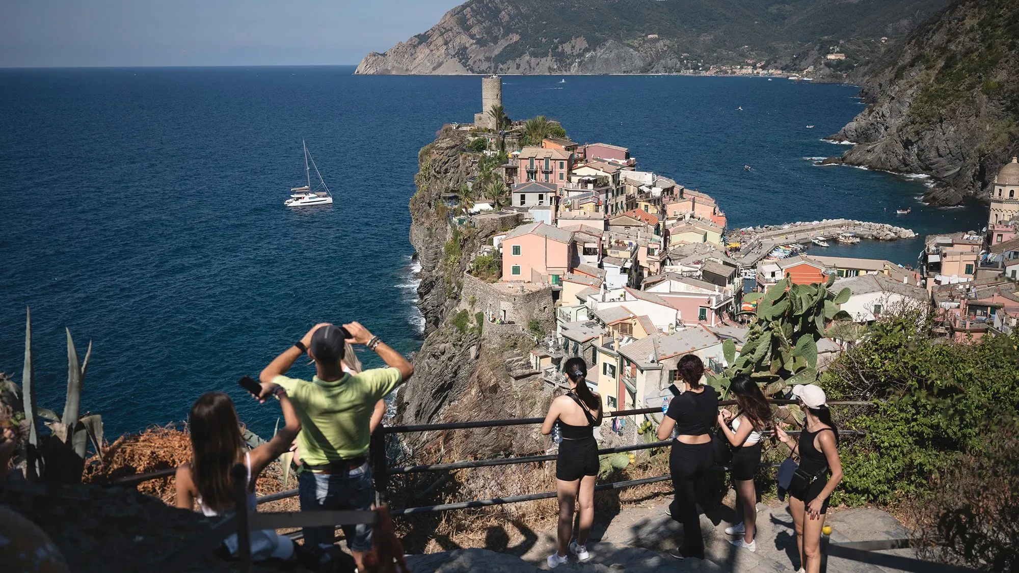 مشهد ساحر لقرية إيطالية ساحلية، حيث يقف زوار على حافة منحدر يلتقطون الصور للمناظر الخلابة للبحر والمنازل الملونة.