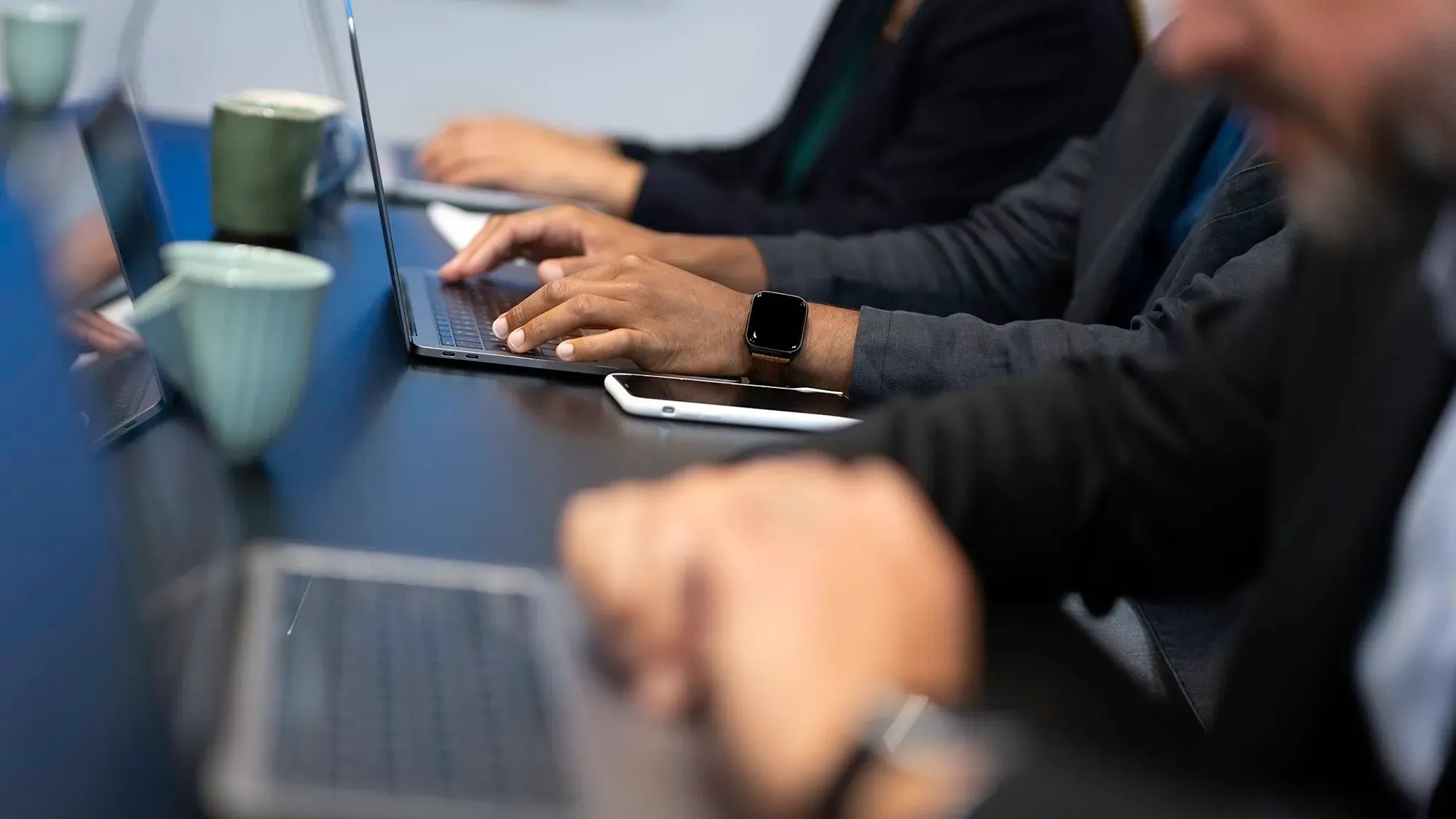 أشخاص يعملون على أجهزة كمبيوتر محمولة في اجتماع، مع أكواب قهوة على الطاولة، مما يعكس زيادة الطلب على وظائف الذكاء الاصطناعي.