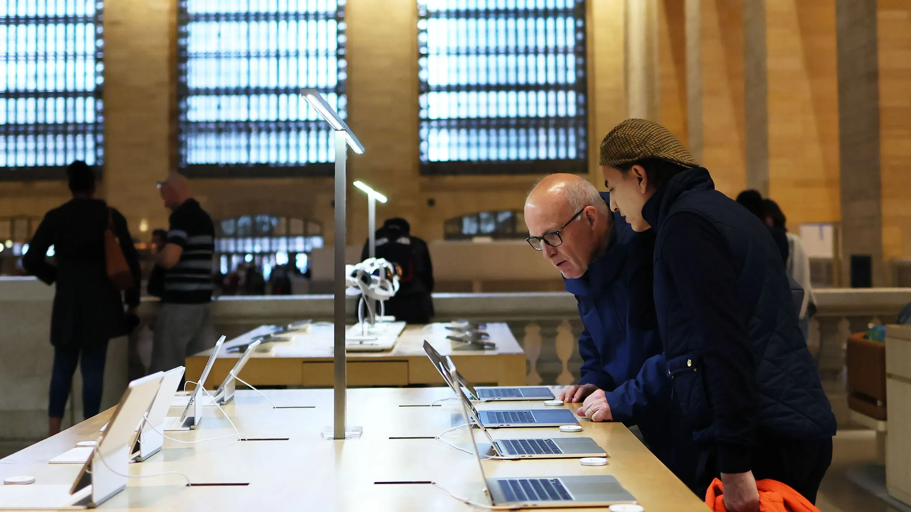 متسوقان يتفقدان أجهزة الكمبيوتر في متجر إلكترونيات، مع وجود عروض للمنتجات تحت ضوء طبيعي من النوافذ الكبيرة، في سياق التعريفات الجمركية.