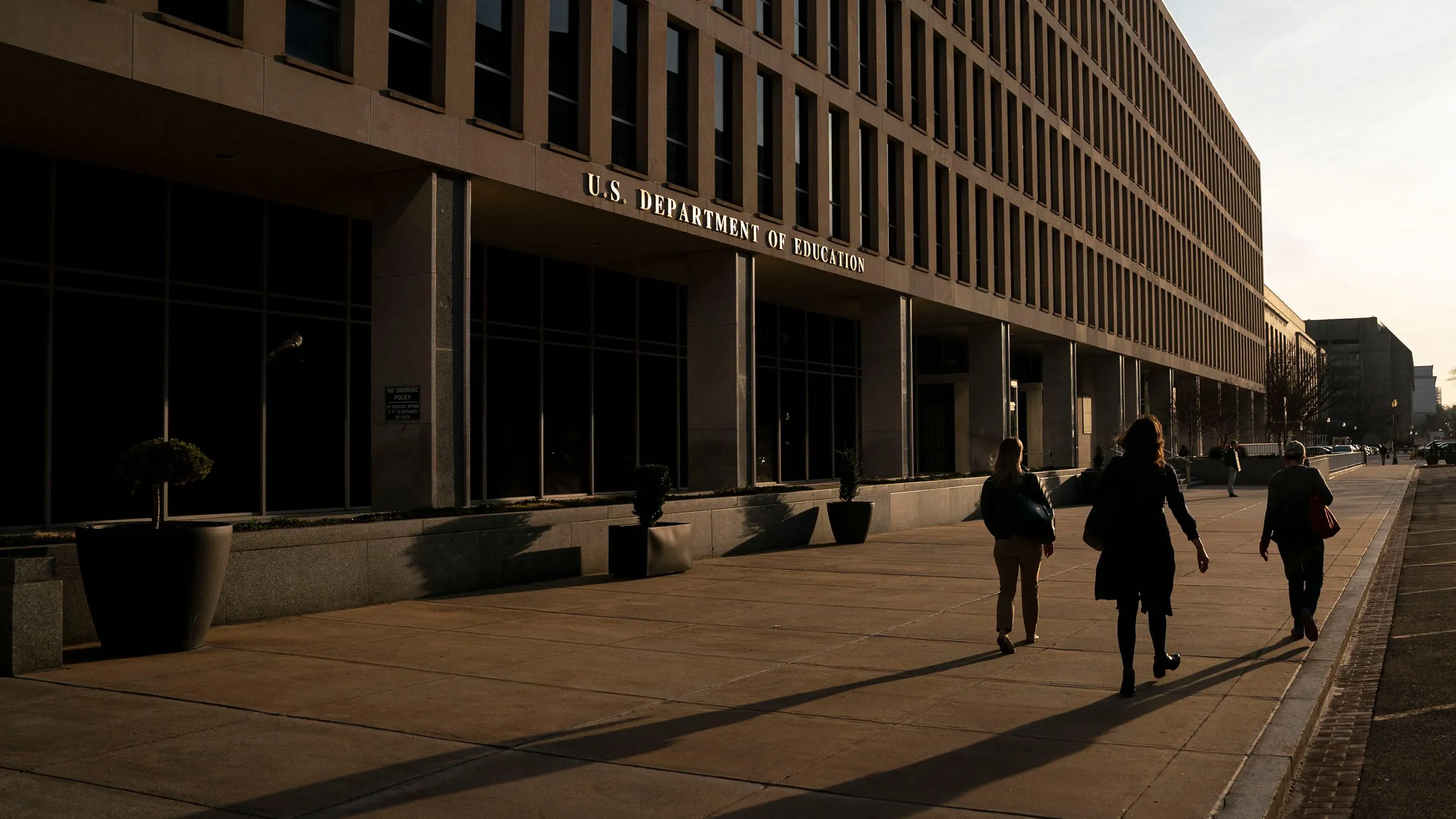 واجهة مبنى وزارة التعليم الأمريكية، حيث يسير عدد من الموظفين في اتجاهات مختلفة، مع وجود أشعة الشمس تضيء المكان.