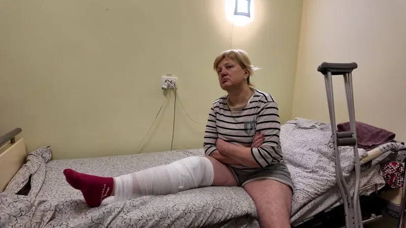 امرأة تجلس على سرير في مستشفى، ترتدي ملابس مريحة وساقها مغطاة برباط طبي، تعكس آثار الحرب في خيرسون.