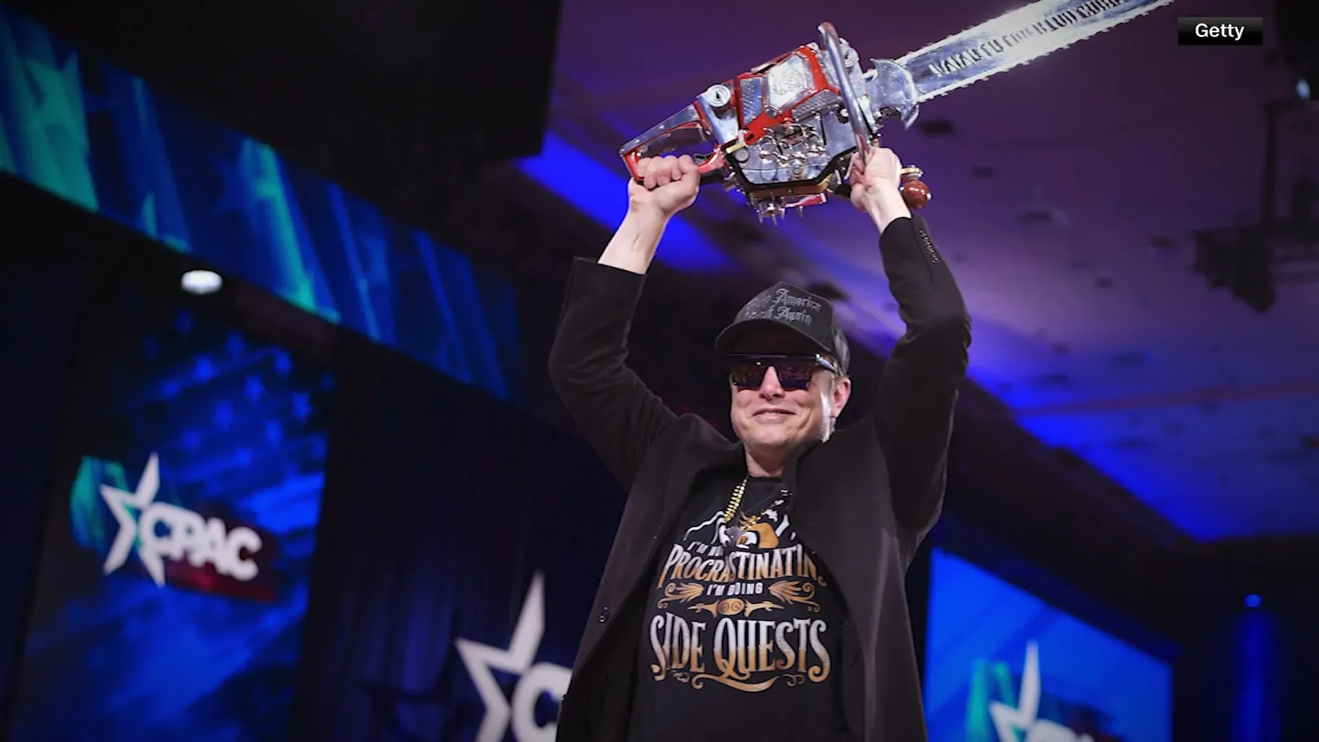 إيلون ماسك يرفع منشاراً كهربائياً في مؤتمر CPAC، مع خلفية ملونة، مما يعكس تأثيره على السياسة والإدارة الفيدرالية.