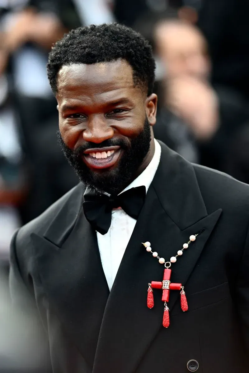 رجل مبتسم يرتدي بدلة رسمية مع قلادة مميزة، يظهر في مهرجان كان السينمائي، مع خلفية من الحضور.