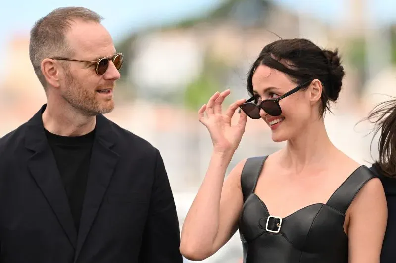 مخرج سينمائي وممثلة يسيران معًا في مهرجان كان، حيث تبرز الأناقة والابتسامة في أجواء احتفالية.