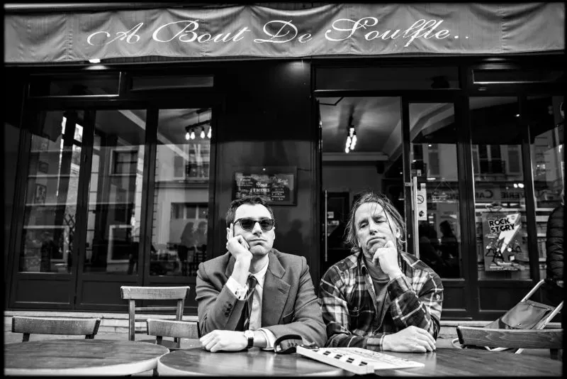 رجلان يجلسان أمام مقهى يحمل لافتة "A Bout De Souffle"، أحدهما يرتدي نظارات شمسية بينما يبدو الآخر غير رسمي.