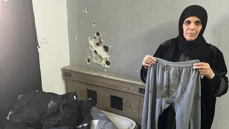 امرأة فلسطينية مسنّة تحمل بنطالًا ممزقًا في غرفة نومها، تظهر على الجدار آثار الرصاص، تعبيرًا عن الفقدان والمعاناة في مخيم بلاطة للاجئين.