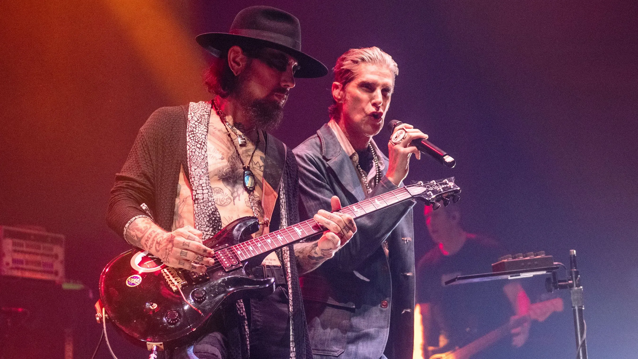 أعضاء فرقة جينز أديكشن يؤدون على المسرح، مع عازف الجيتار ديف نافارو والمغني بيري فاريل، وسط أضواء ملونة، في عرض متوتر.