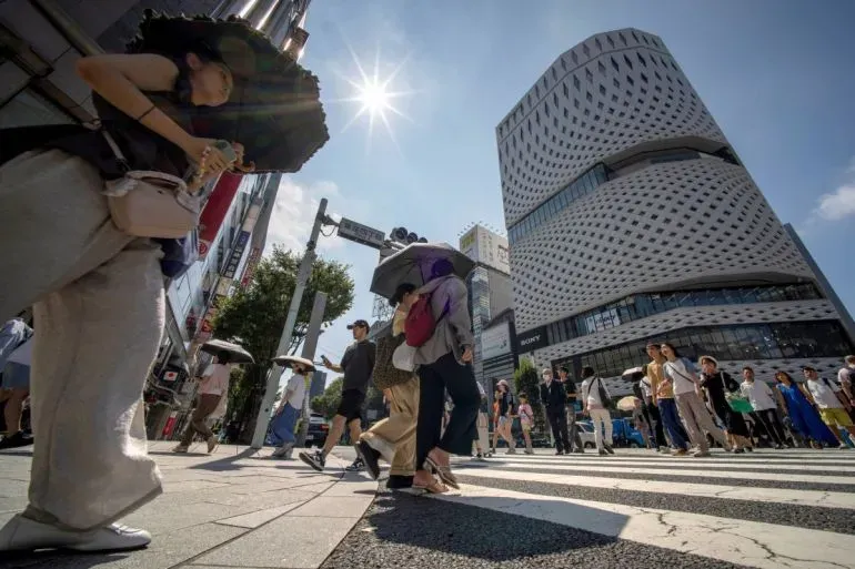 أشخاص يتجولون في شوارع مدينة يابانية تحت أشعة الشمس الساطعة، مع استخدام المظلات للحماية من الحرارة الشديدة.