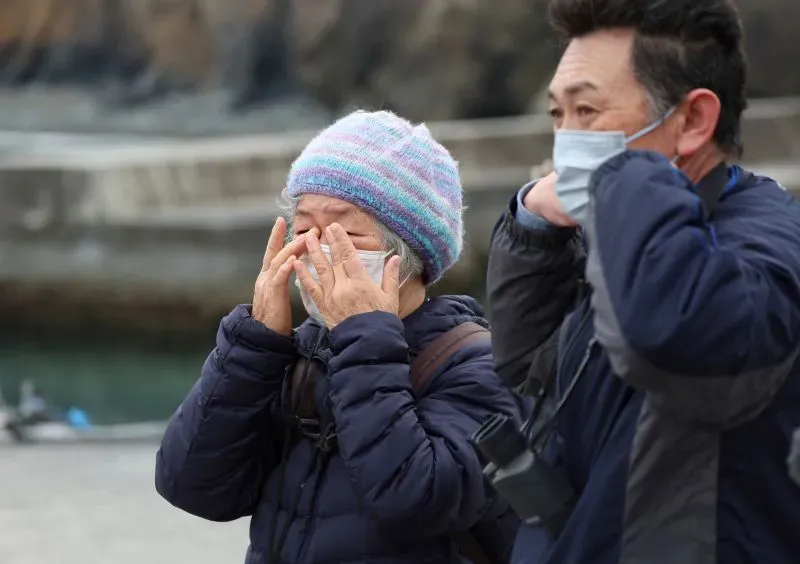 سيدة مسنّة ترتدي قبعة ملونة وتغطي وجهها بيدها، بينما يقف رجل بجانبها، يعبران عن مشاعر الحزن بعد حريق الغابات في أوفوناتو.