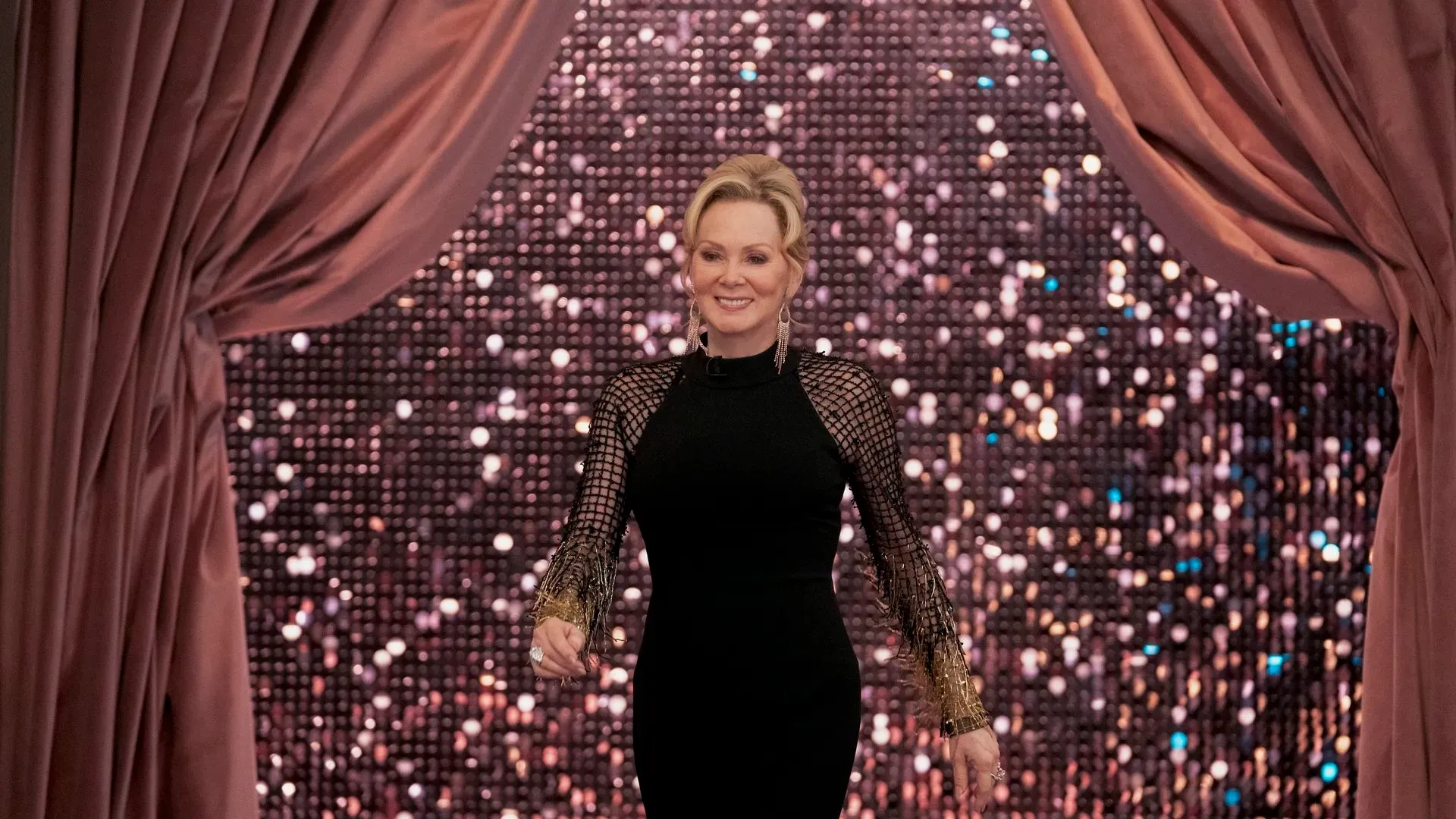 جين سمارت ترتدي فستانًا أسود أنيقًا وتبتسم أثناء ظهورها على المسرح، محاطة بأضواء براقة، مما يعكس أجواء عرضها في برودواي.