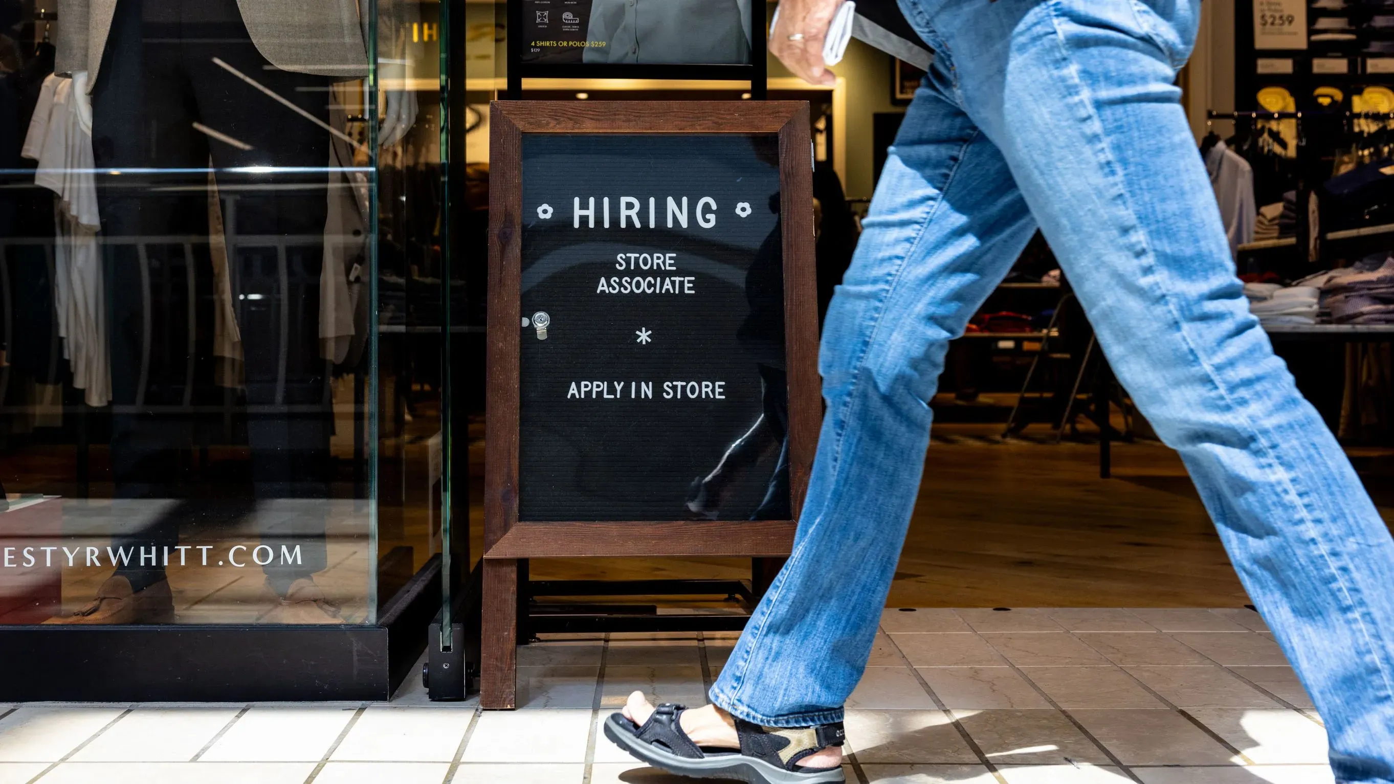 لافتة "نحن نوظف" خارج متجر، مع شخص يرتدي جينز يمشي بجانبها، تعكس زيادة فرص العمل في قطاع التجزئة.