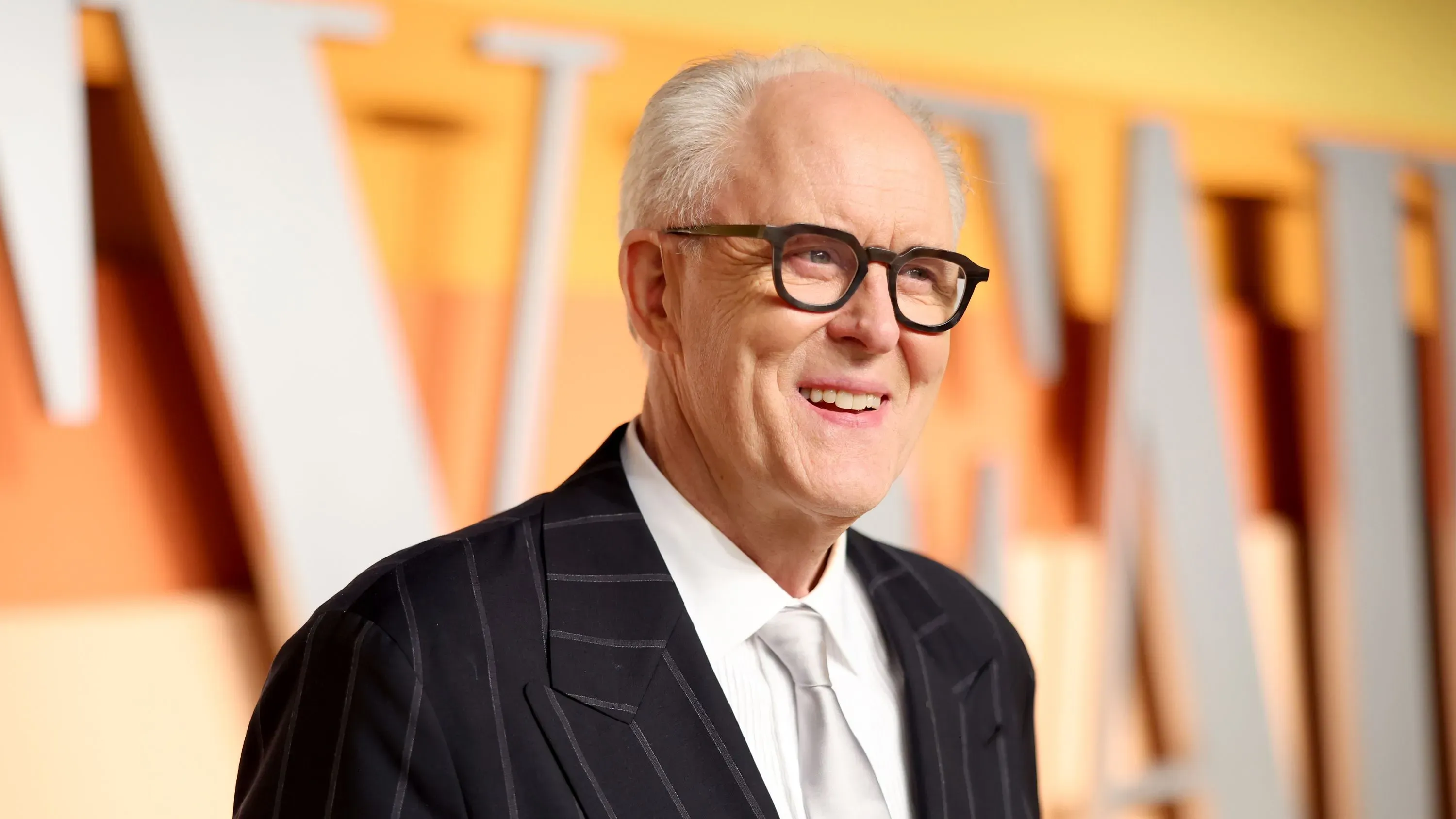 جون ليثغو مبتسم، يرتدي بدلة أنيقة مع نظارات، يتحدث عن ذكرياته في مسلسل "الصخرة الثالثة من الشمس" وتأثيره على مسيرته الفنية.