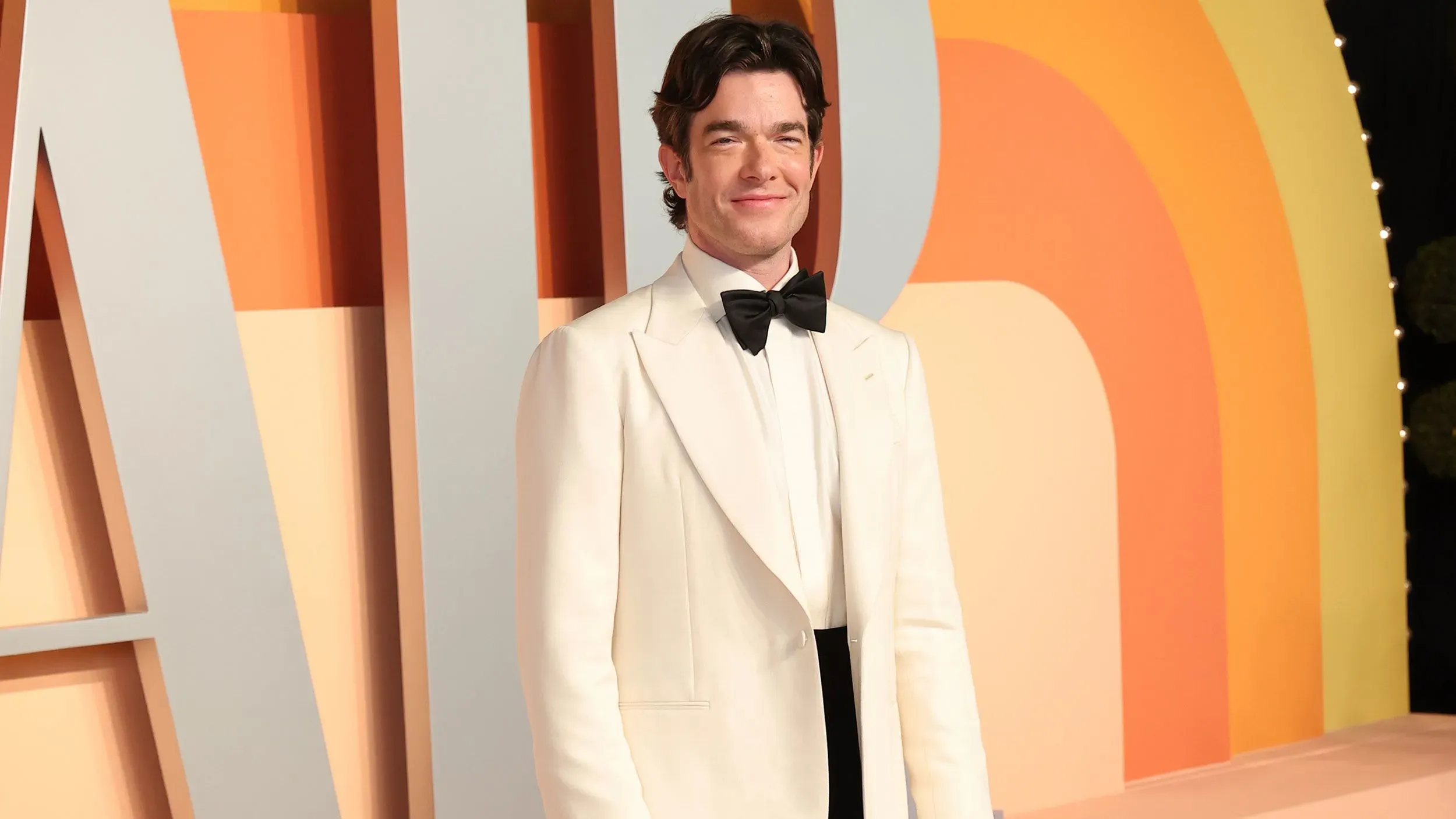 جون مولاني يرتدي بدلة بيضاء مع ربطة عنق سوداء، مبتسمًا أمام خلفية ملونة، في حدث يبرز حضوره كأب وكوميدي.