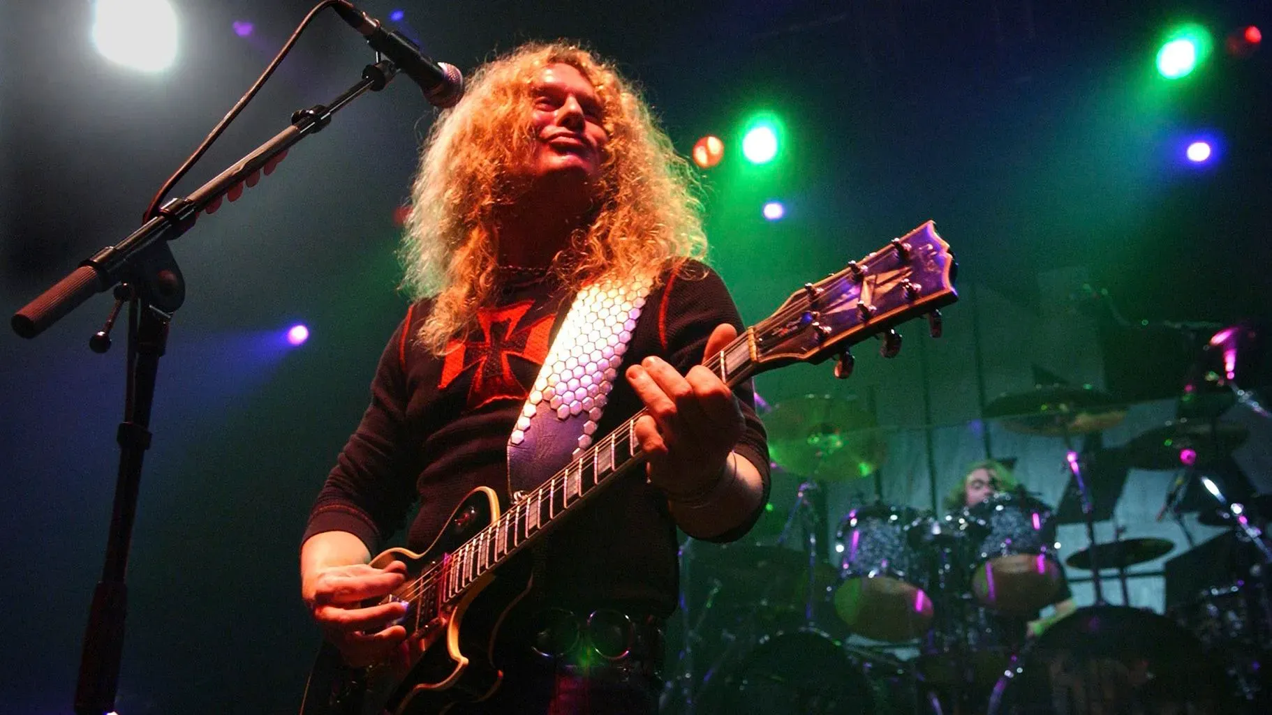 عازف الجيتار جون سايكس يؤدي على المسرح، محاطًا بأضواء ملونة، مع التركيز على موهبته الموسيقية وتأثيره في عالم الروك.