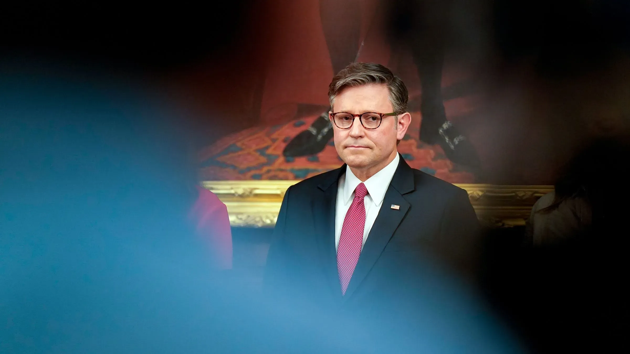 مايك جونسون، رئيس مجلس النواب، يقف في قاعة المجلس مع تعبير جاد، بينما خلفه يظهر رسم تاريخي، وسط أجواء من التوتر السياسي.