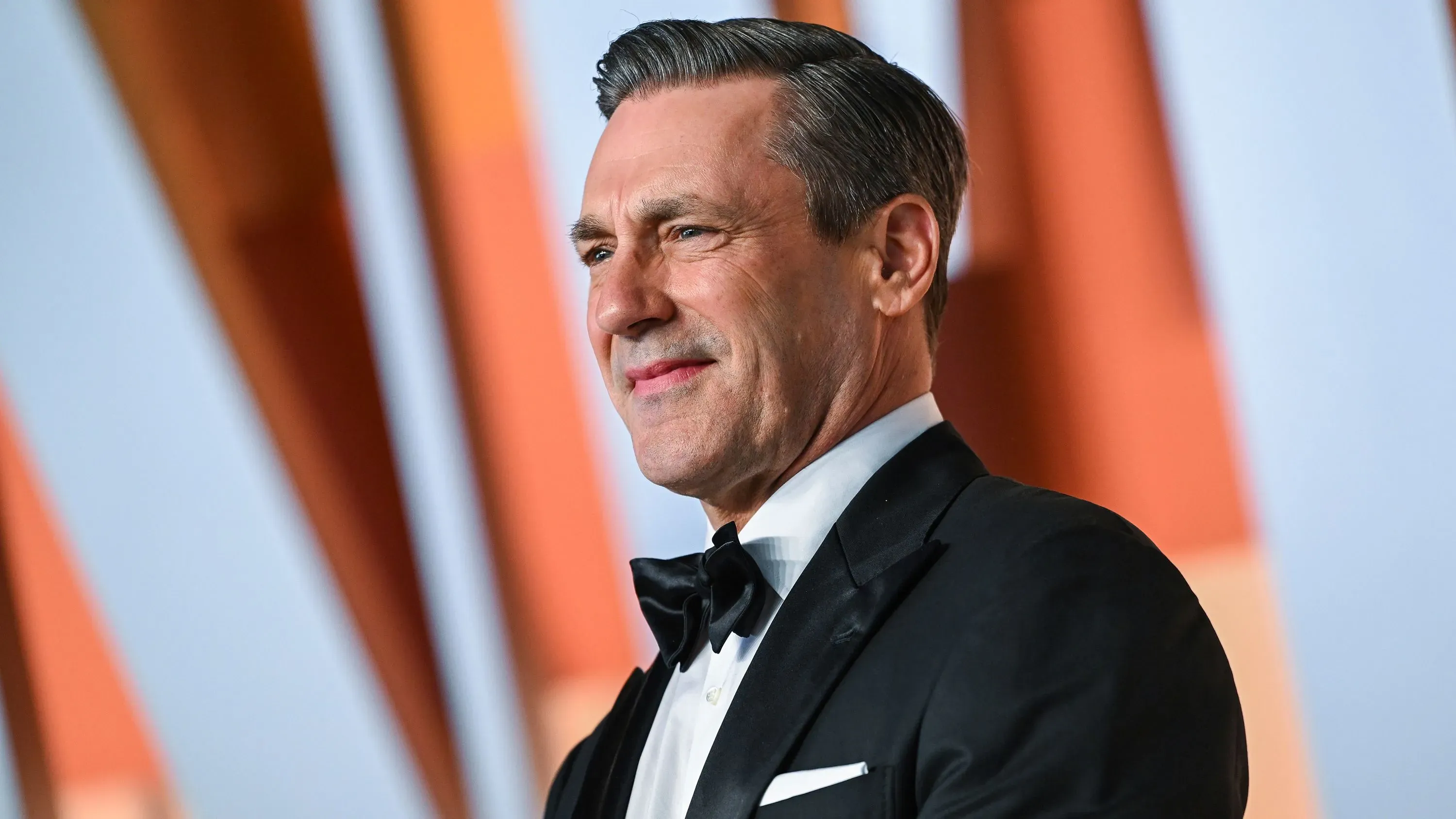 جون هام مبتسم في بدلة رسمية، يظهر في حدث عام، مع خلفية ملونة تعكس أجواء احتفالية، مرتبطًا بمسلسل "The Big Fix".