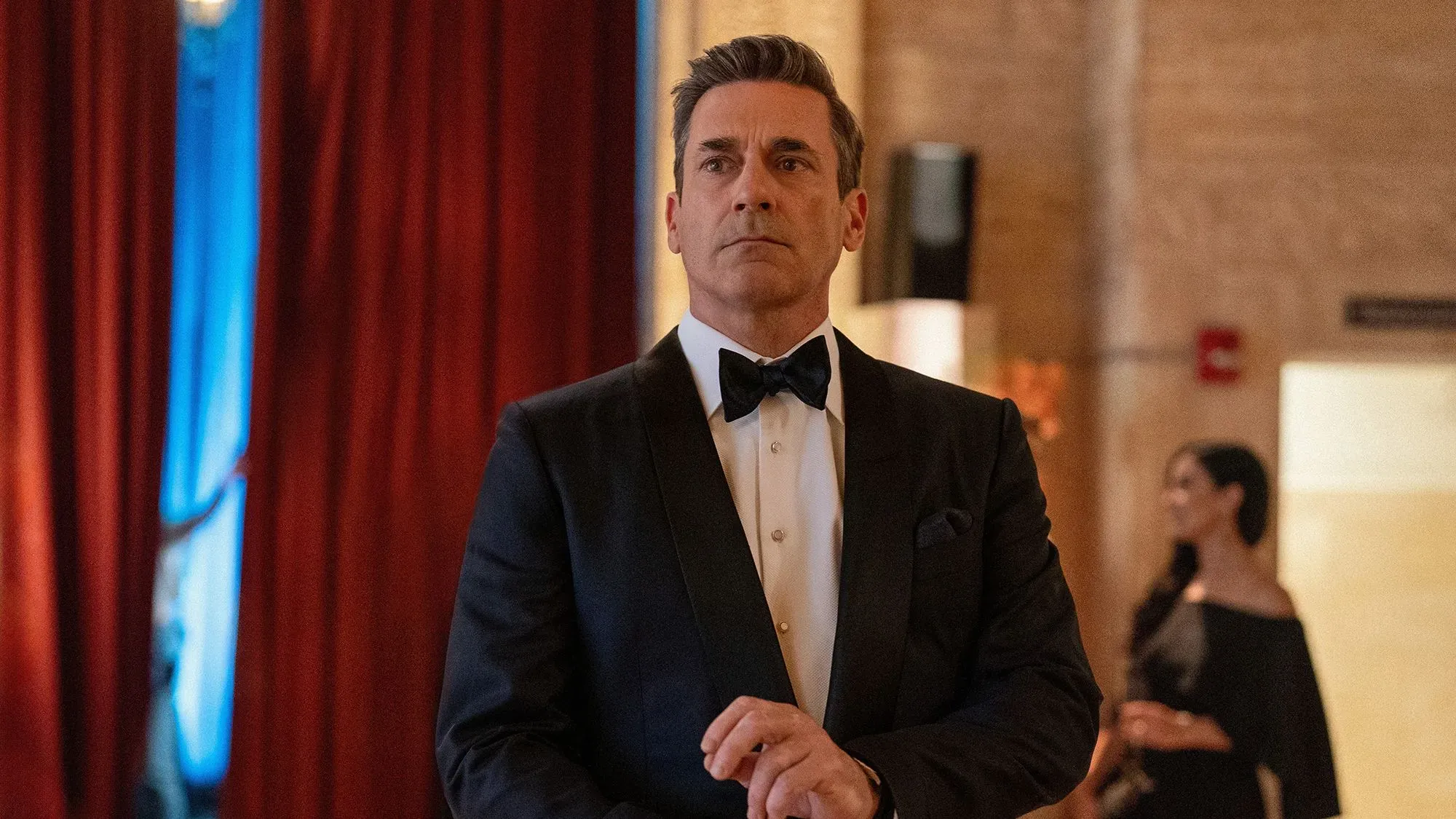 شخص يرتدي بدلة رسمية مع ربطة عنق، يبدو عليه القلق في حدث اجتماعي راقٍ، مع خلفية من الستائر الحمراء.