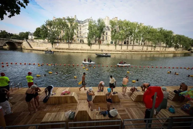 سباحون يستعدون للسباحة في نهر السين بباريس، مع وجود طوافات حمراء تحدد مناطق السباحة، وأبنية تاريخية في الخلفية.