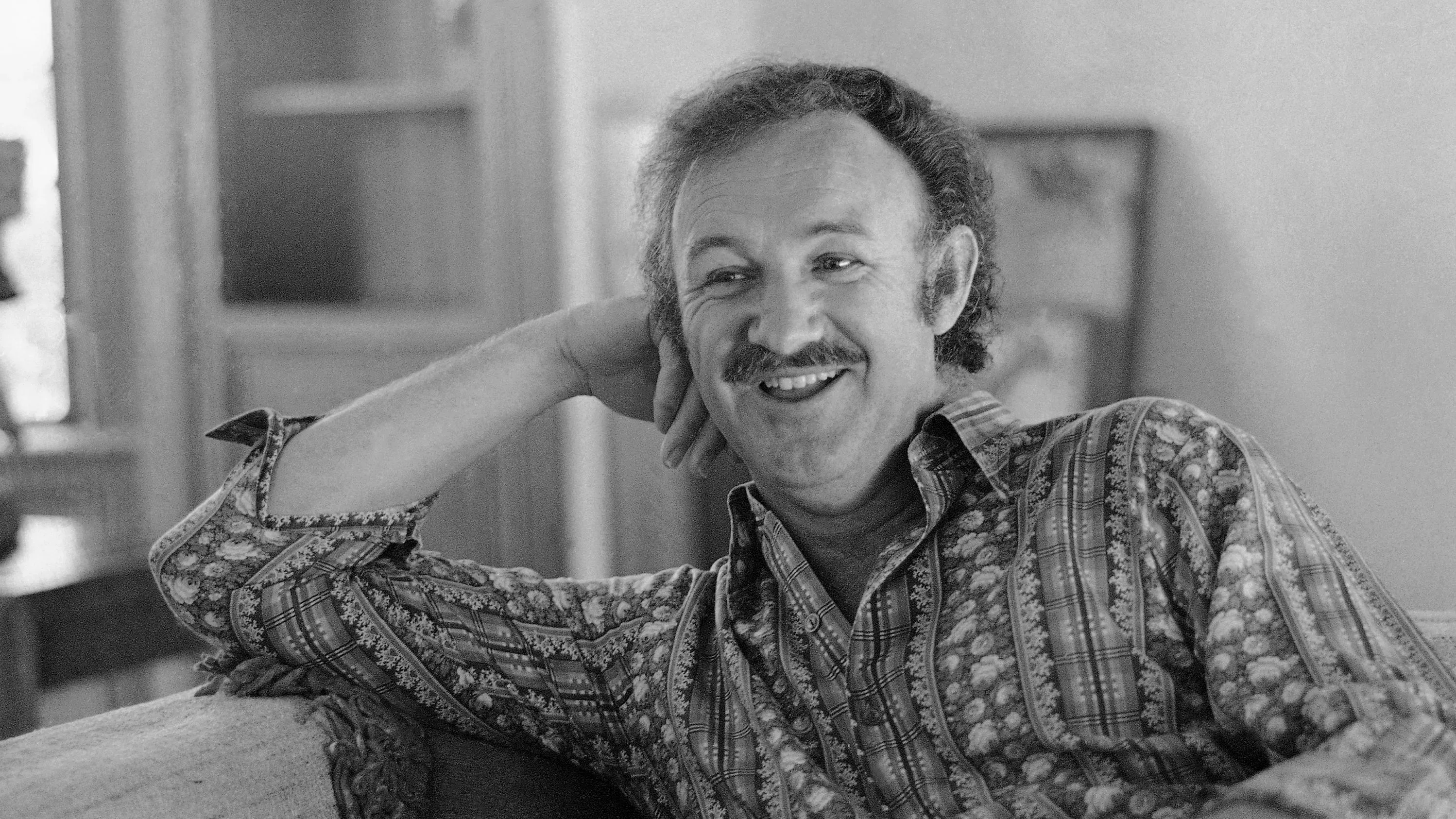 صورة بالأبيض والأسود لجين هاكمان مبتسمًا، يجلس على أريكة مع ذراعه ممدودة، تعكس شخصيته الودية.