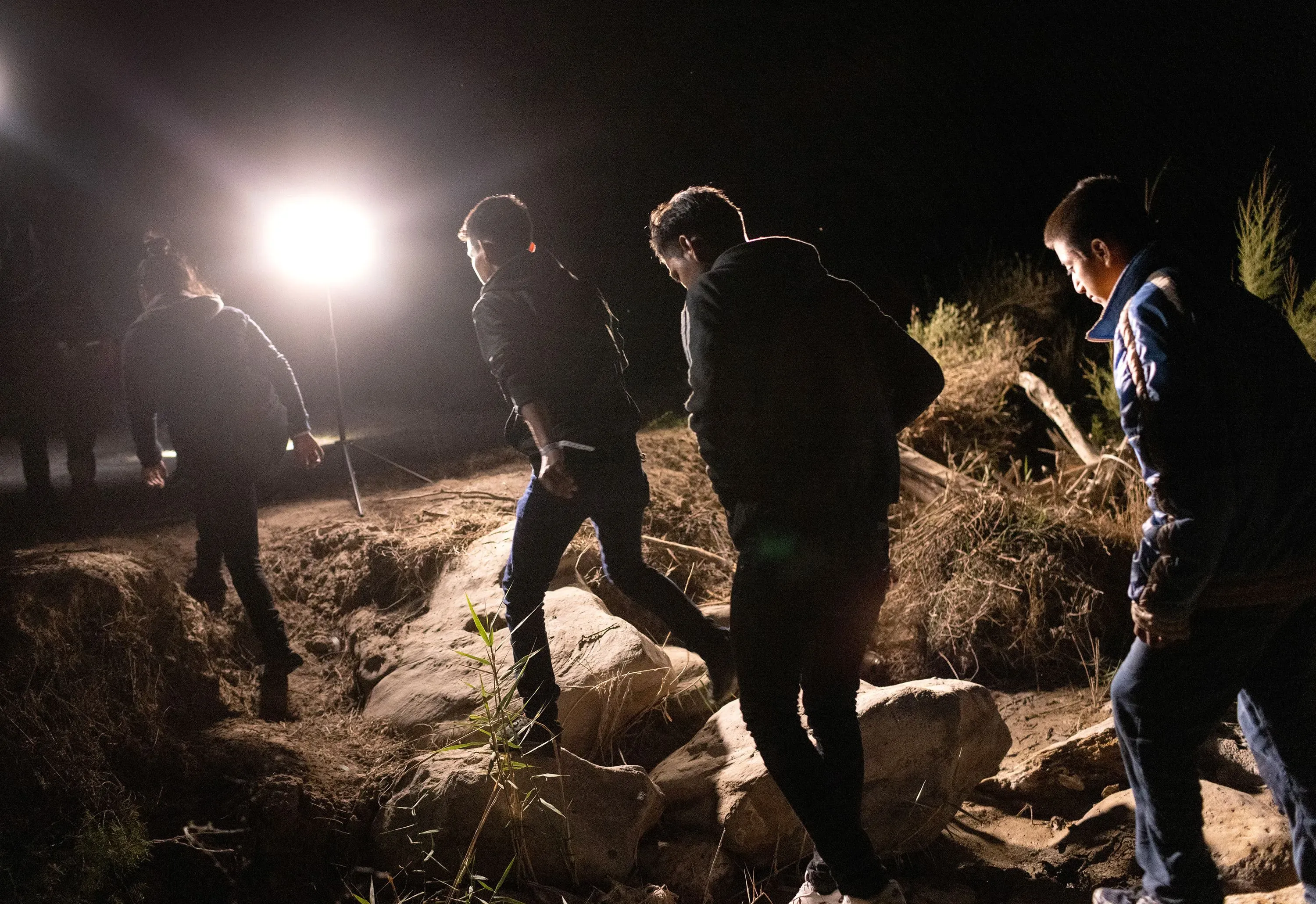 مجموعة من الشباب الغواتيماليين يسيرون في الظلام نحو الحدود، مع وجود أضواء ساطعة في الخلفية، تعكس محاولاتهم للهجرة.