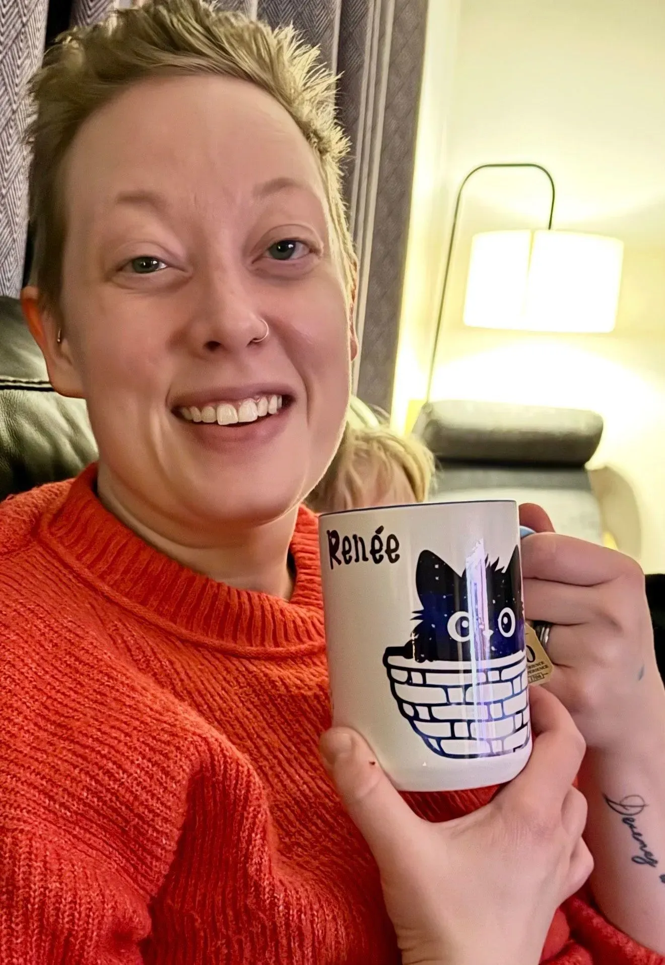 امرأة مبتسمة ترتدي سترة حمراء، تحمل فنجانًا مزينًا بصورة قطة، في خلفية منزلية دافئة. تعكس الصورة أجواء عائلية مريحة.