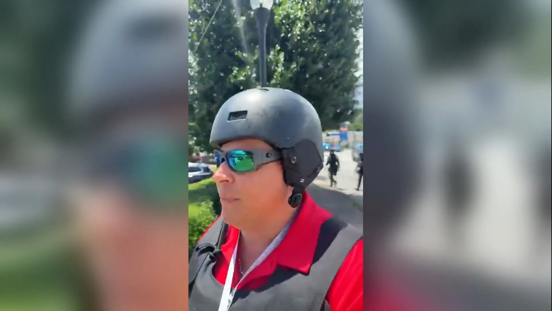 ماريو جيفارا، الصحفي المحتجز، يرتدي سترة واقية ونظارات شمسية خلال تغطيته لمظاهرة في أتلانتا، مع وجود قوات إنفاذ القانون في الخلفية.