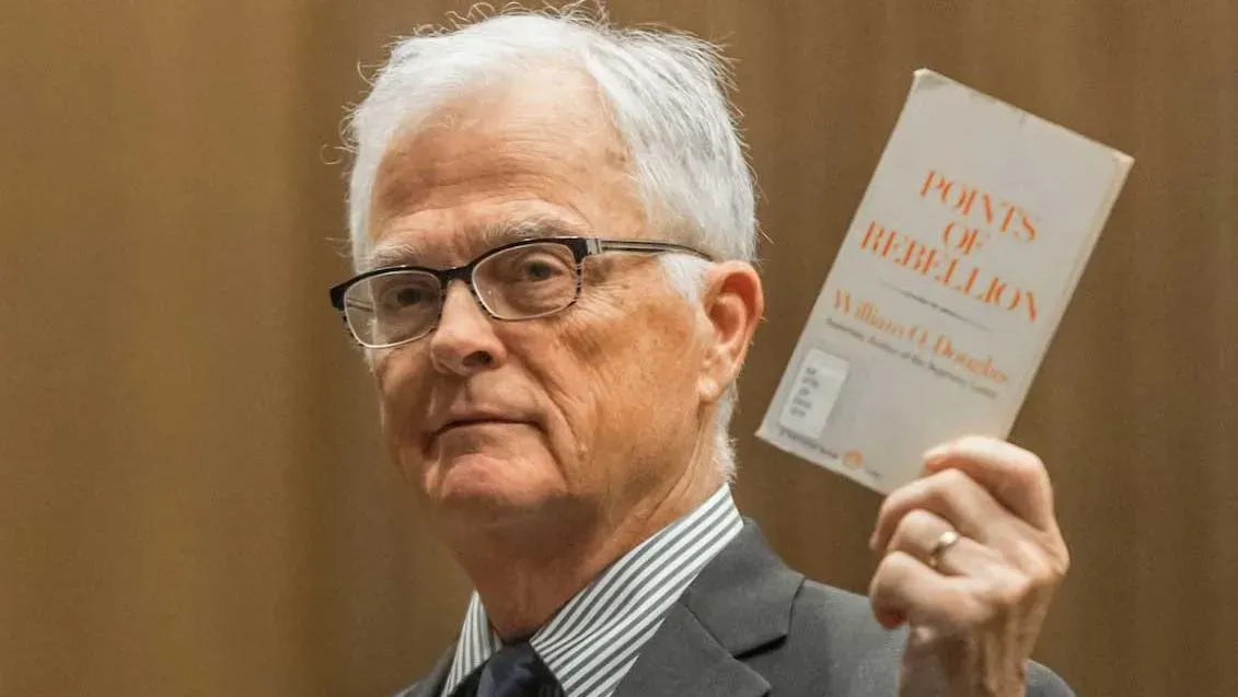 رجل مسن يرتدي نظارات، يحمل كتاب "نقاط من التمرد" في جلسة قانونية تتعلق بفصل الموظفين الفيدراليين.