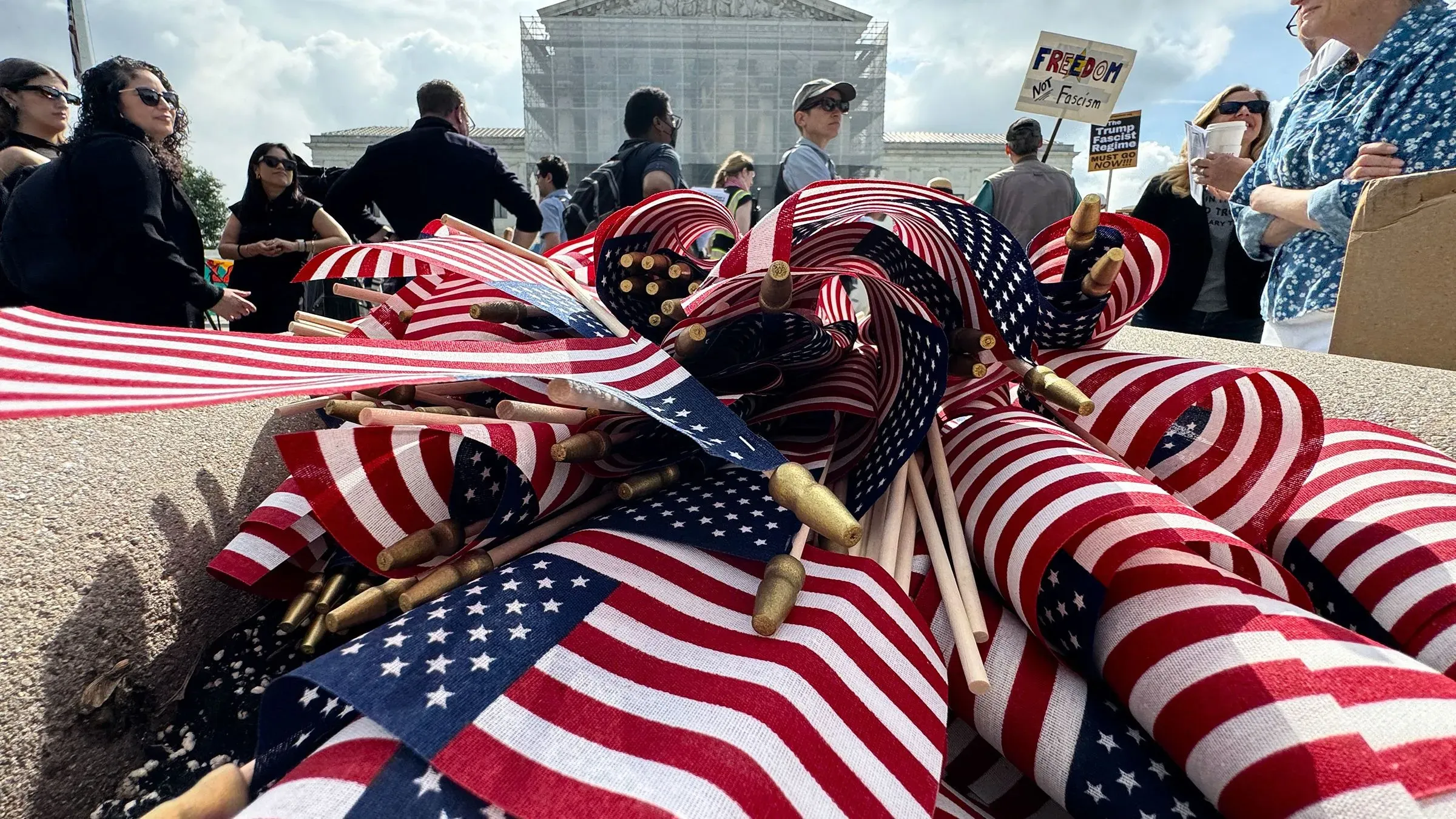 تظهر الصورة مجموعة من الأعلام الأمريكية ملتفة حول عصي، مع وجود متظاهرين في الخلفية يحملون لافتات تعبر عن آرائهم حول سياسة ترامب المتعلقة بالمواطنة.
