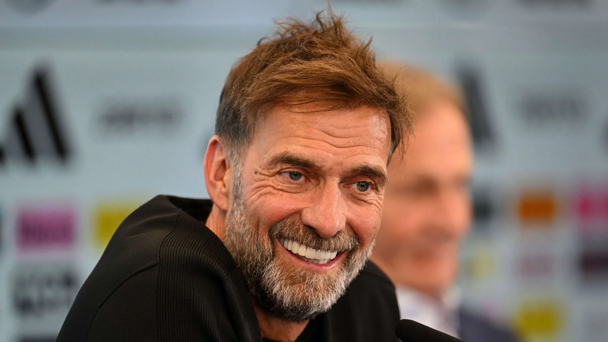 يورغن كلوب يبتسم خلال مؤتمر صحفي بعد تعيينه مدرباً جديداً للمنتخب الألماني لكرة القدم، مع التركيز على دوره في بناء الفريق واستمرارية المنظومة.