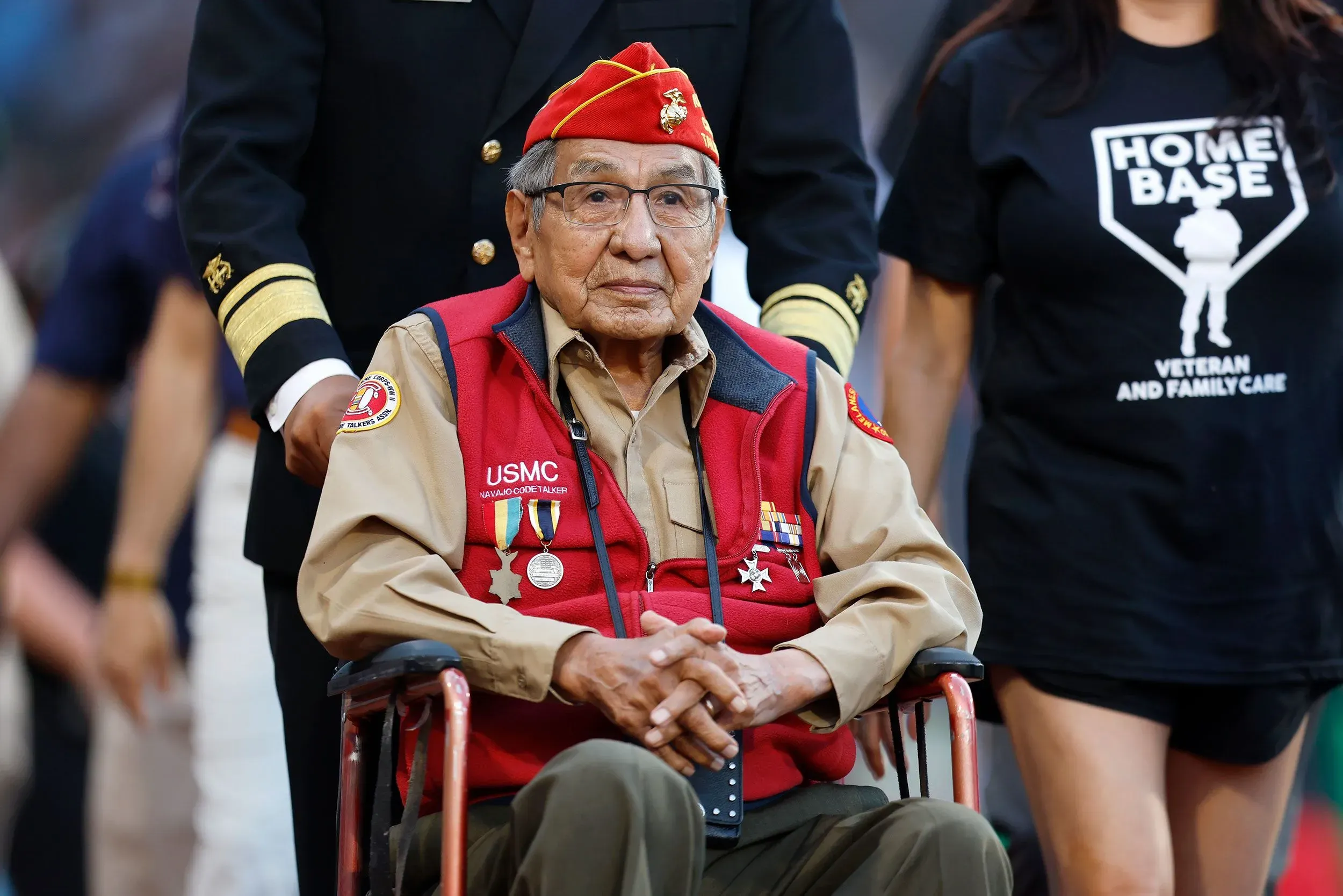 رجل مسن يرتدي زيًا عسكريًا مع وساماته، يجلس في كرسي متحرك أثناء احتفال تكريمي، يرمز إلى إسهامات المتحدثين بشفرة النافاجو في الحرب العالمية الثانية.
