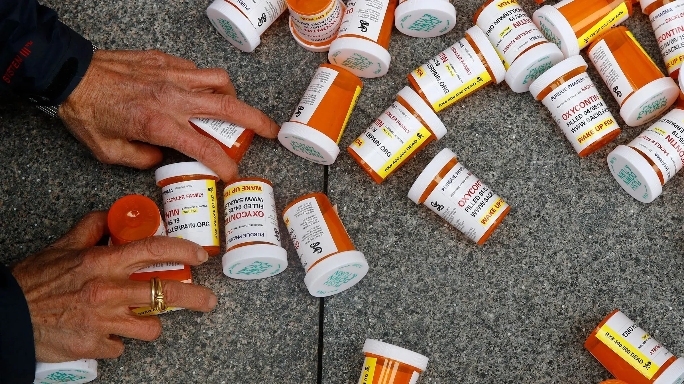 زجاجات دواء فارغة مكدسة على الأرض، بينما تمسك يد بشرية ببعضها، مما يسلط الضوء على أزمة المواد الأفيونية وتأثيرها على المجتمع.