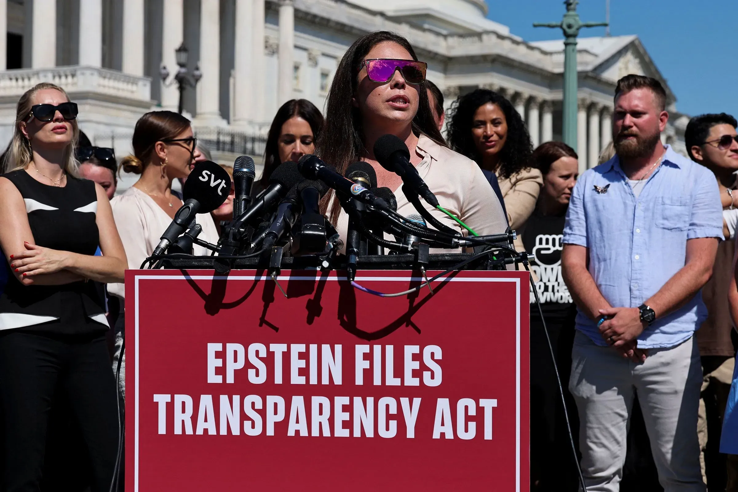 امرأة تتحدث أمام ميكروفونات في تجمع لدعم قانون الشفافية في ملفات إبشتاين، مع مجموعة من المتظاهرين خلفها.