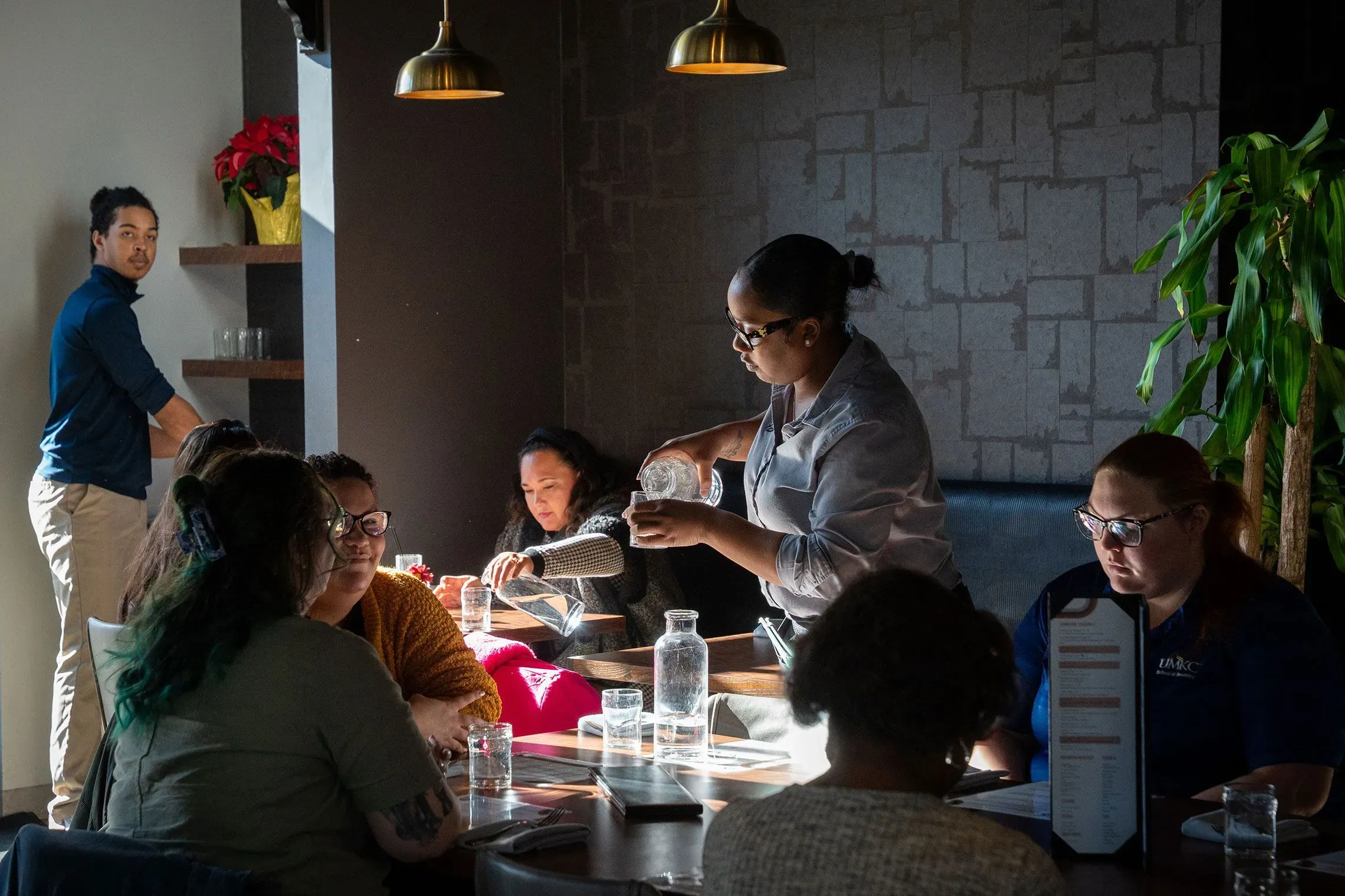مجموعة من الأشخاص يجلسون في مطعم، وموظفة تقدم الماء لطاولة تضم زبائن، مع وجود نباتات في الخلفية.
