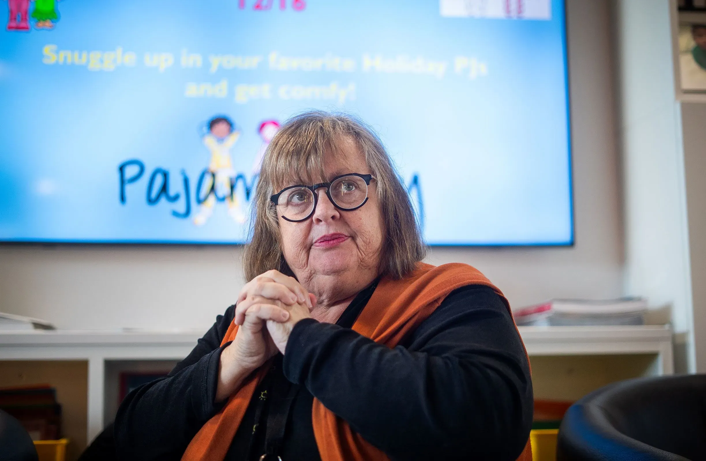 امرأة ترتدي نظارات وقميصًا أسود مع سترة برتقالية، تجلس في غرفة مليئة بالألعاب، مع شاشة خلفها تعرض عبارة "استعدوا للاحتفال".