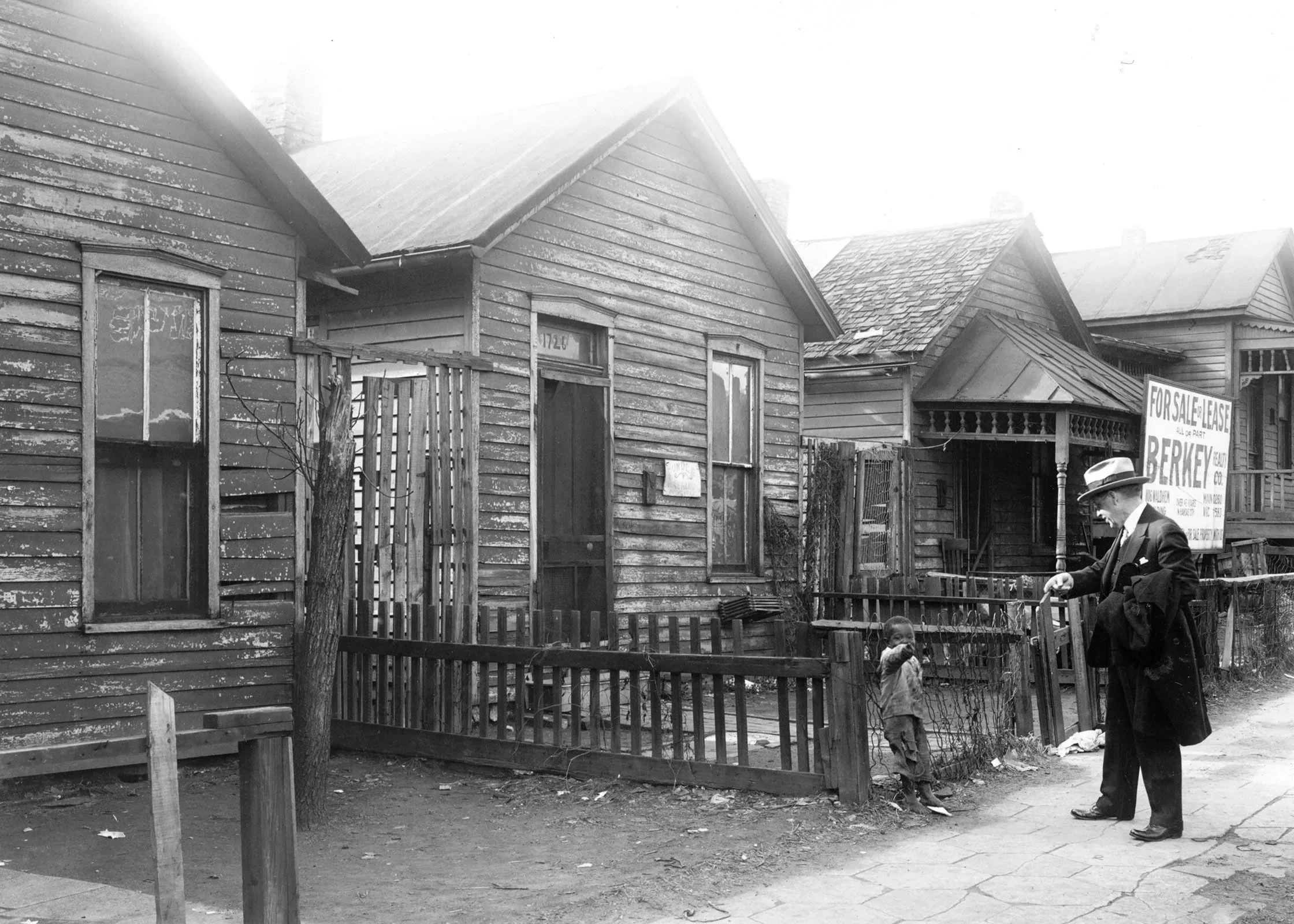 صورة تاريخية تظهر رجلًا يرتدي بدلة وقبعة يتحدث مع طفل صغير بالقرب من منازل قديمة في مدينة كانساس سيتي، تعكس آثار الفصل العنصري.