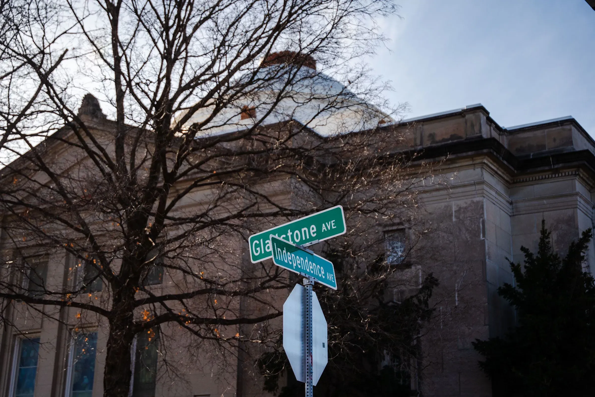 تظهر الصورة لافتة تشير إلى تقاطع شارعي غلاستون واستقلال، مع خلفية لمبنى تاريخي وأشجار عارية، تعكس تاريخ الفصل العنصري في كانساس سيتي.