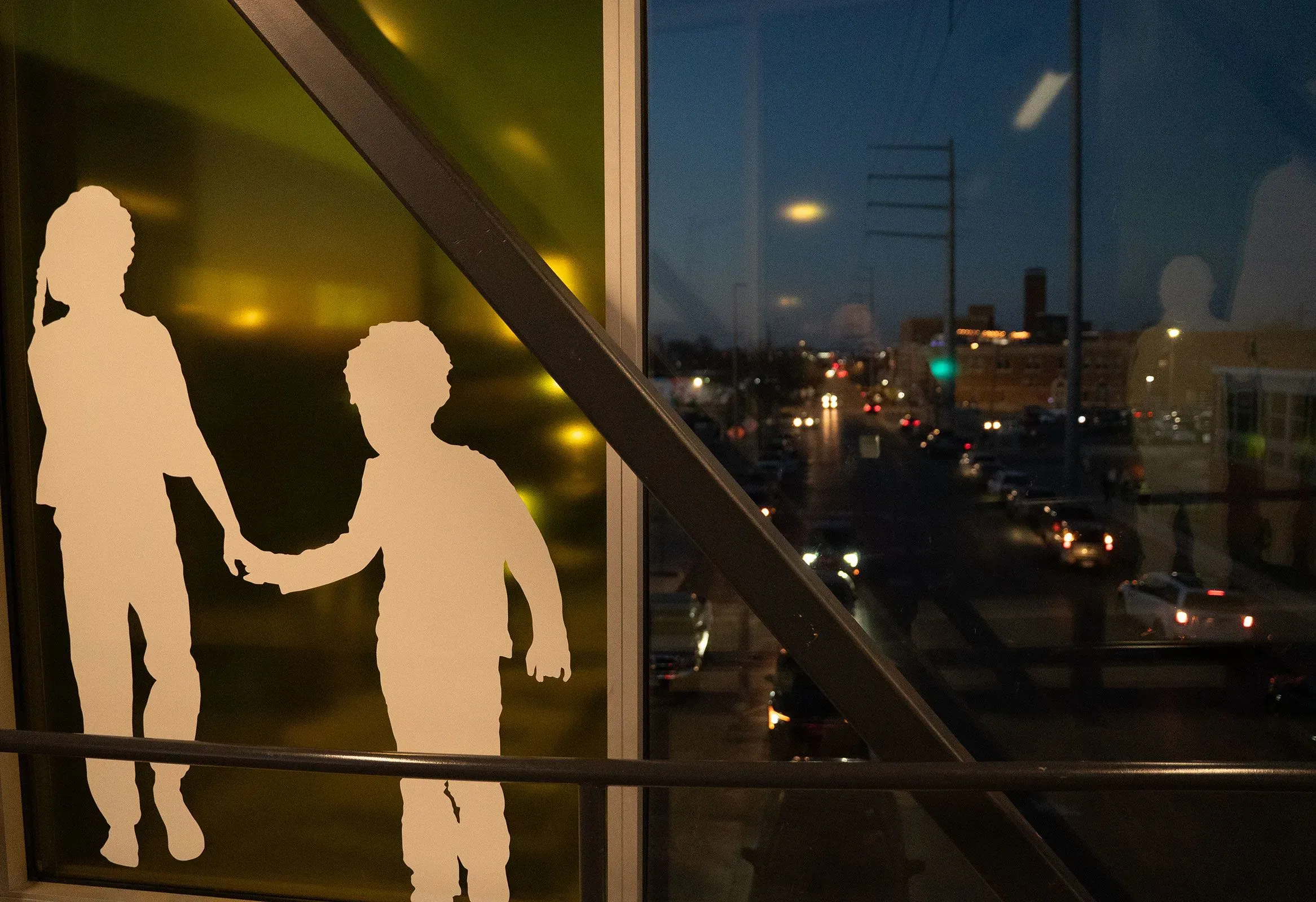 صورة توضح silhouettes لطفلين يمسكان بأيديهما، خلف زجاج نافذة تطل على شارع مزدحم في مدينة كانساس سيتي عند الغسق.