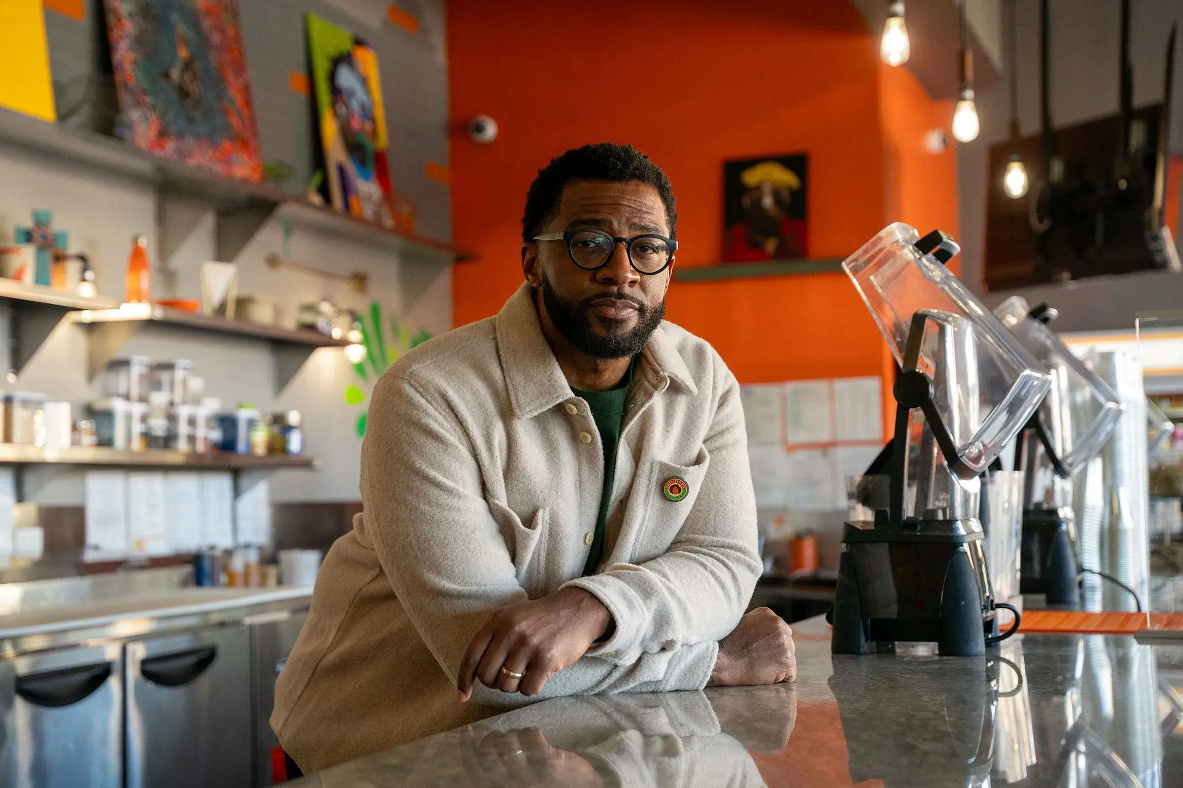 رجل أسود يرتدي نظارات يقف أمام منضدة في مقهى، مع خلفية ملونة تحتوي على لوحات فنية، يعكس روح مجتمع كانساس سيتي.