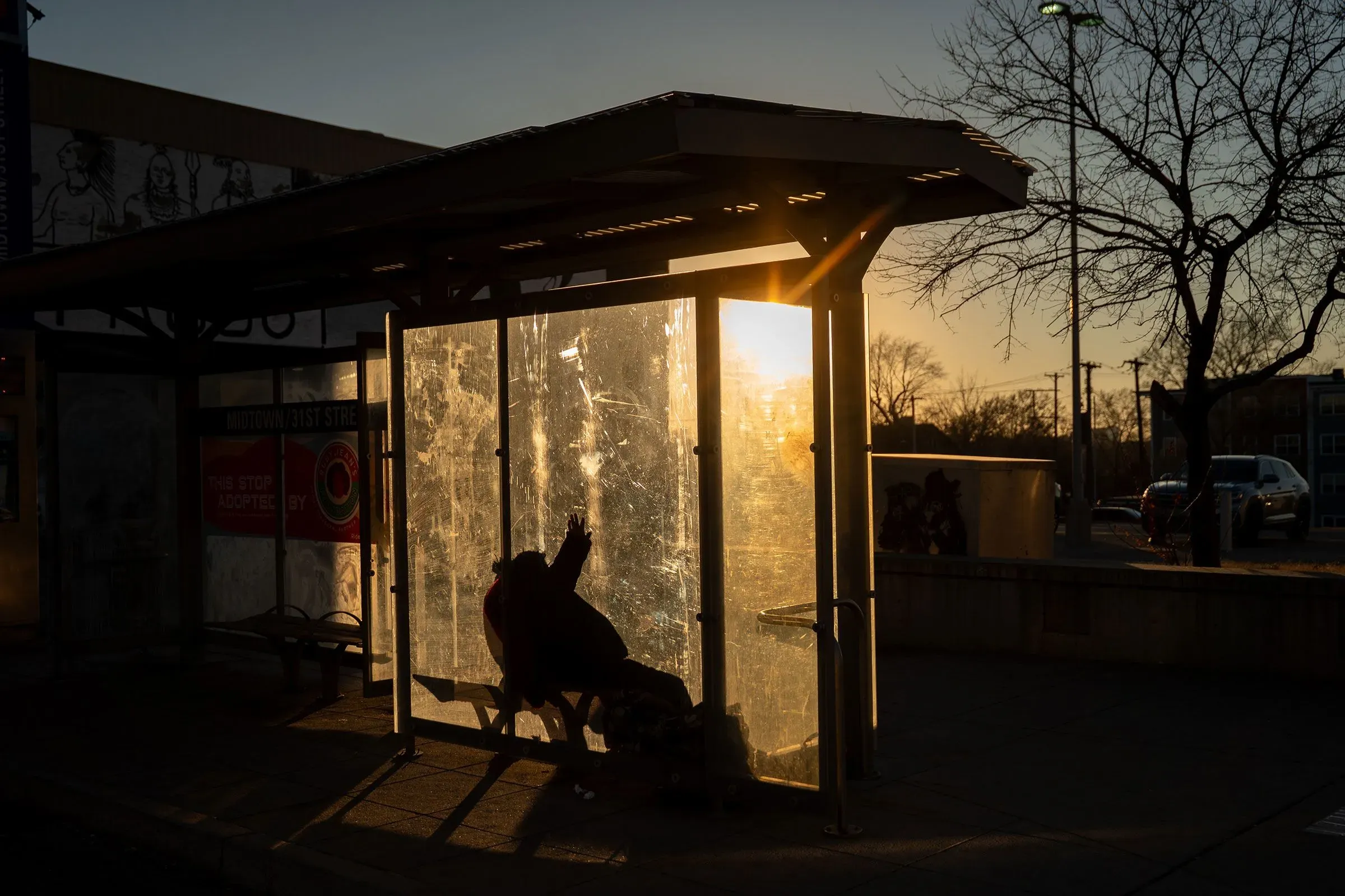 موقف حافلات في مدينة كانساس سيتي، يظهر شخصًا يجلس في الظل بينما تغرب الشمس، مما يعكس الفجوة الاقتصادية والاجتماعية في المنطقة.